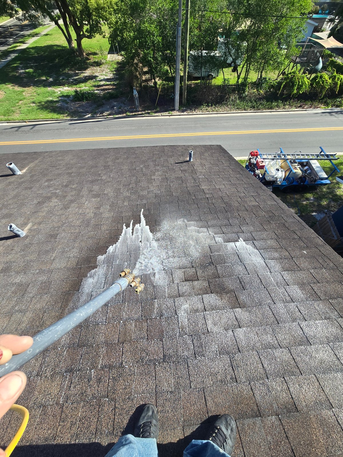 Aerial view of roof cleaning in progress with worker spraying asphalt shingles and equipment visible below on the yard and street.