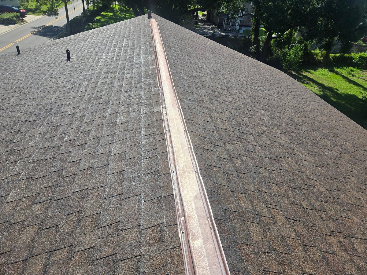 Aerial view of a gray asphalt shingle roof with a metal ridge cap running down the center, surrounded by green trees and residential structures