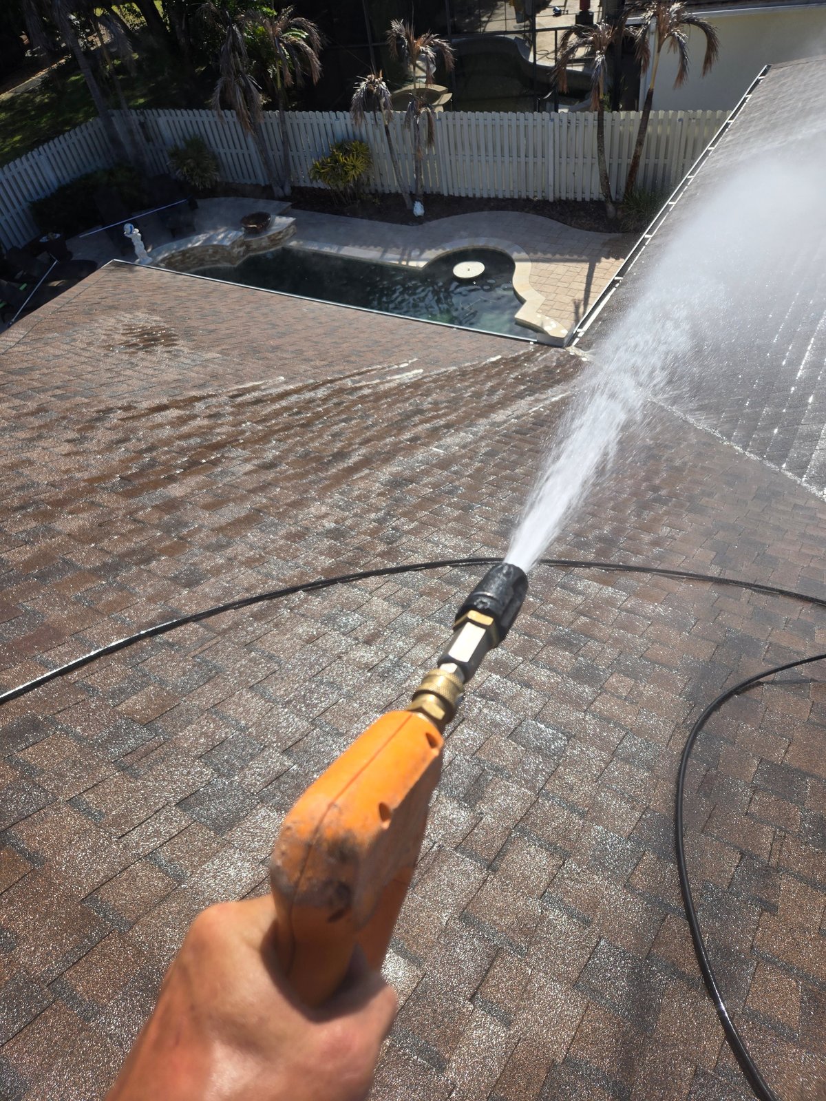 Hand holding pressure washer nozzle spraying water on brick patio with pool and white fence in background