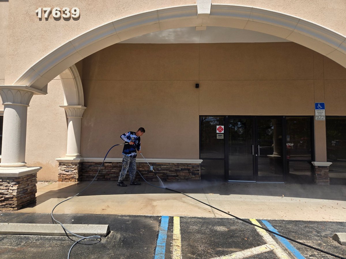 Worker pressure washing the concrete patio of a beige building with arched entryway and dark entrance doors