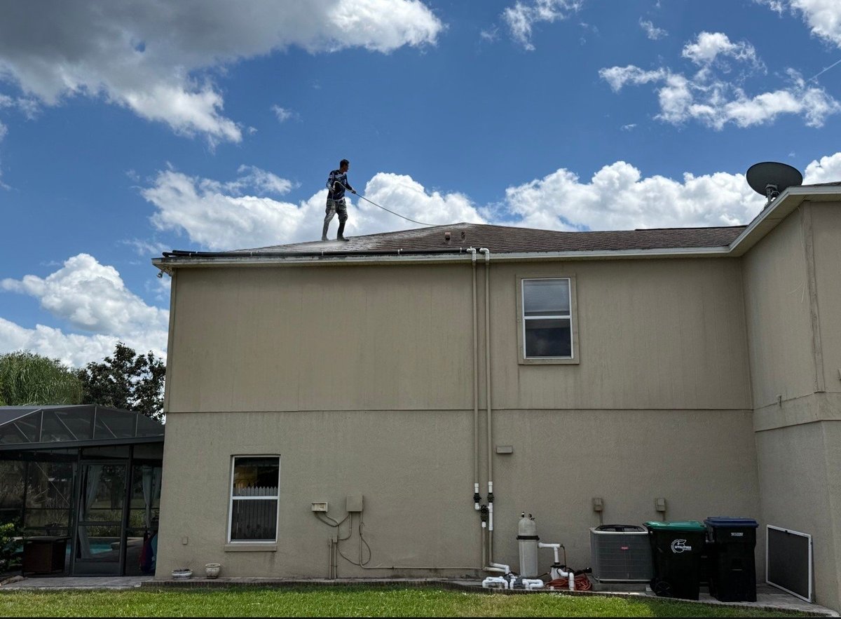 Person standing on the roof of a beige two-story house with a green lawn and blue sky with clouds