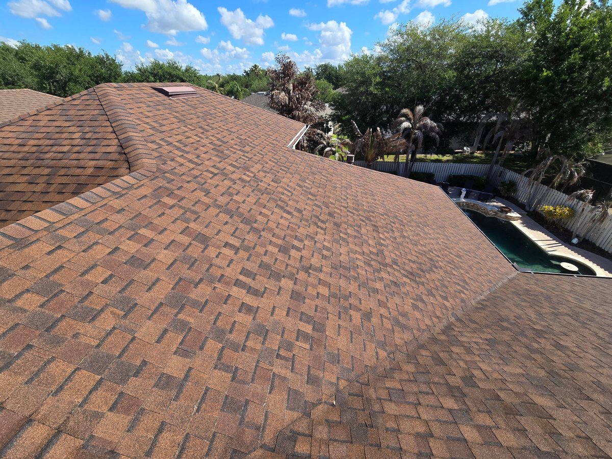 Aerial view of a shingled roof with residential landscape, trees, and a pool visible in the background