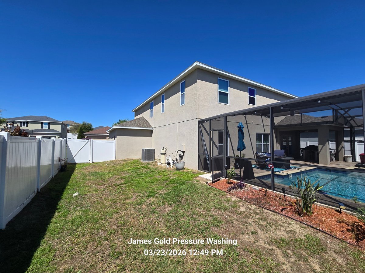 Backyard of two-story beige house with green lawn, enclosed pool area, and clean white fence under clear blue sky