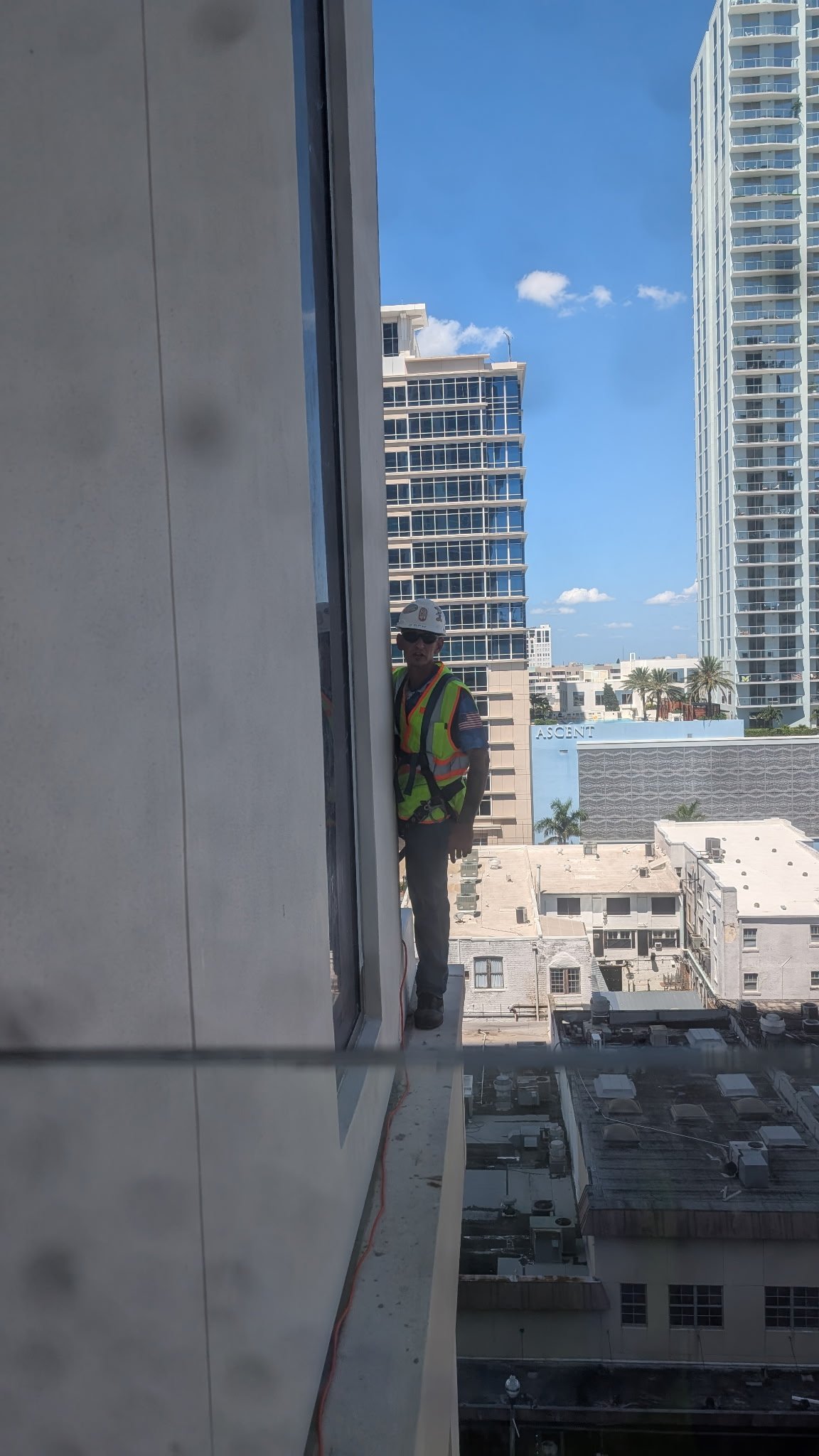 Construction worker in safety vest on high-rise building ledge overlooking urban cityscape with modern buildings and clear sky