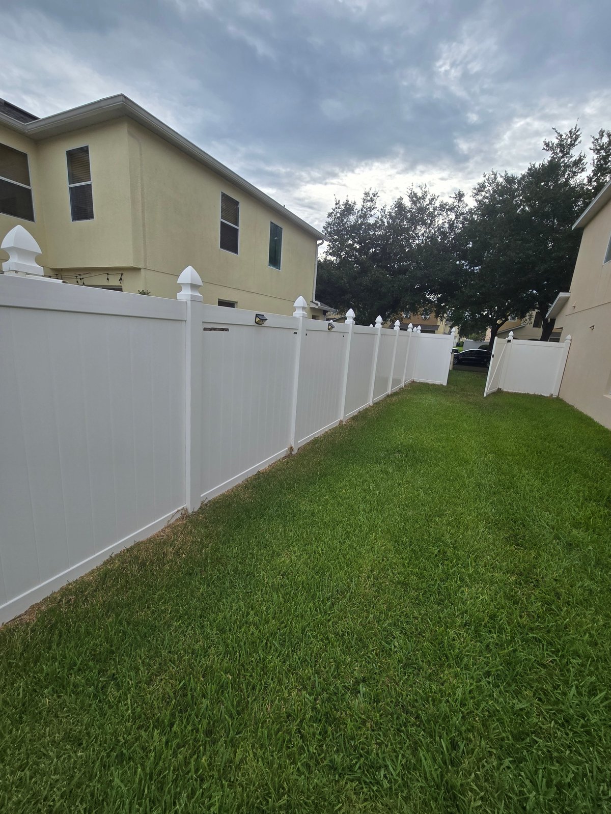 White vinyl fence lining a green lawn beside a yellow two-story house under a cloudy sky