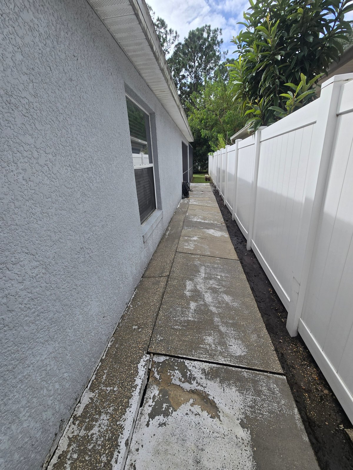 Clean paved side walkway between a light blue stucco house and a white vinyl fence after pressure washing