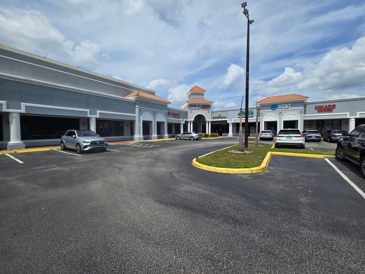 Exterior of a strip mall shopping center with white columns, terra cotta tile roofing, and parked cars in the paved lot