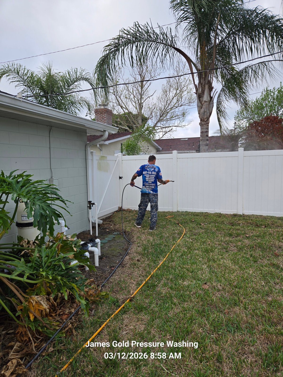 Worker pressure washing residential backyard lawn and pathway next to white fence and garage