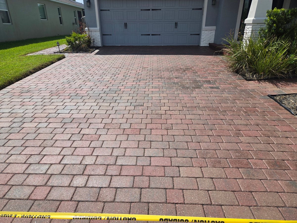 Red brick paver driveway in front of gray garage with yellow caution tape and landscaping on sides