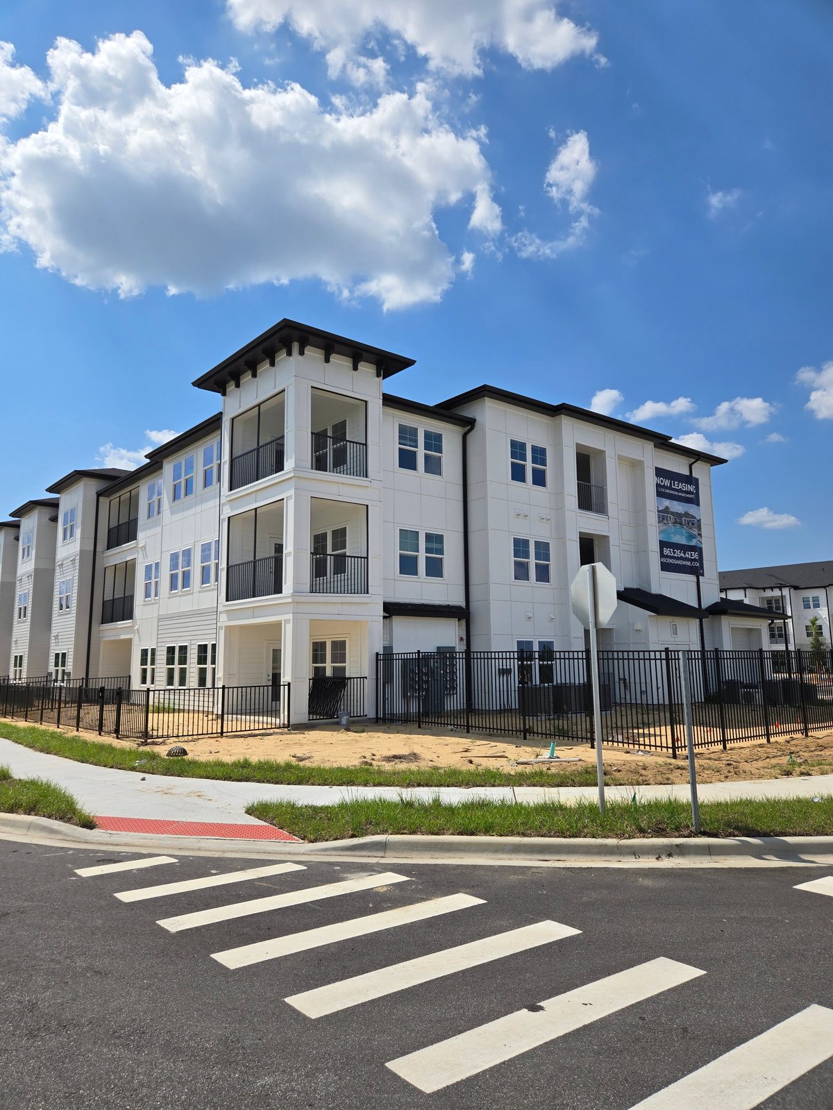 Modern three-story apartment building with white and black exterior, black fencing, and crosswalk in foreground under blue sky with clouds