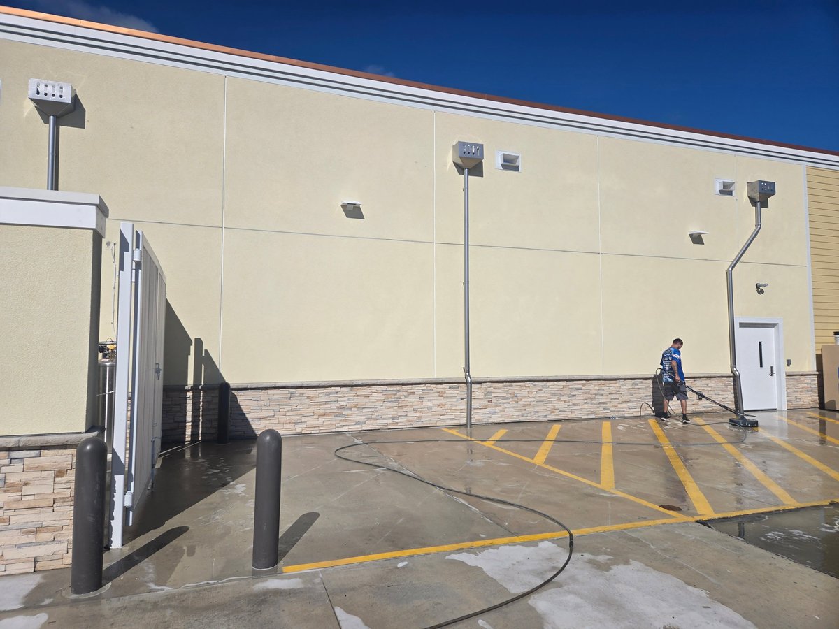 Exterior of large commercial building with tan metal walls, loading dock, parking area with yellow line markings, tall flood lights, and one person visible on the concrete ground.