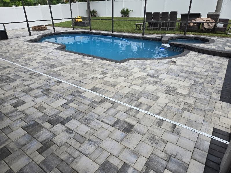 Blue swimming pool with paved deck enclosed by black safety fence, outdoor lounge chairs visible in background