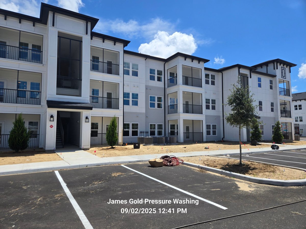 Multi-story apartment complex with white and gray facade under blue sky, featuring parking lot and landscaping.