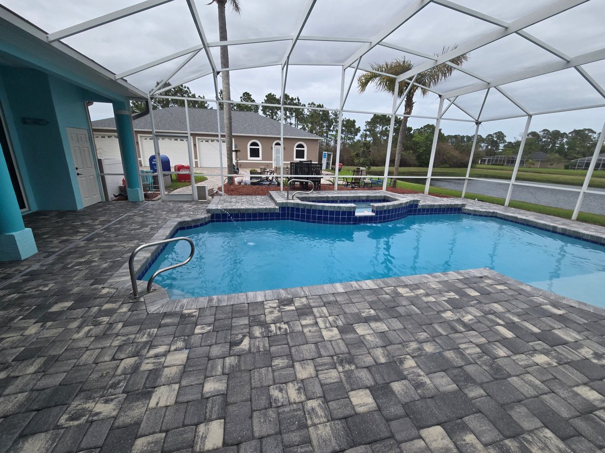 Indoor swimming pool with blue water under screened enclosure, brick patio, and residential buildings in background