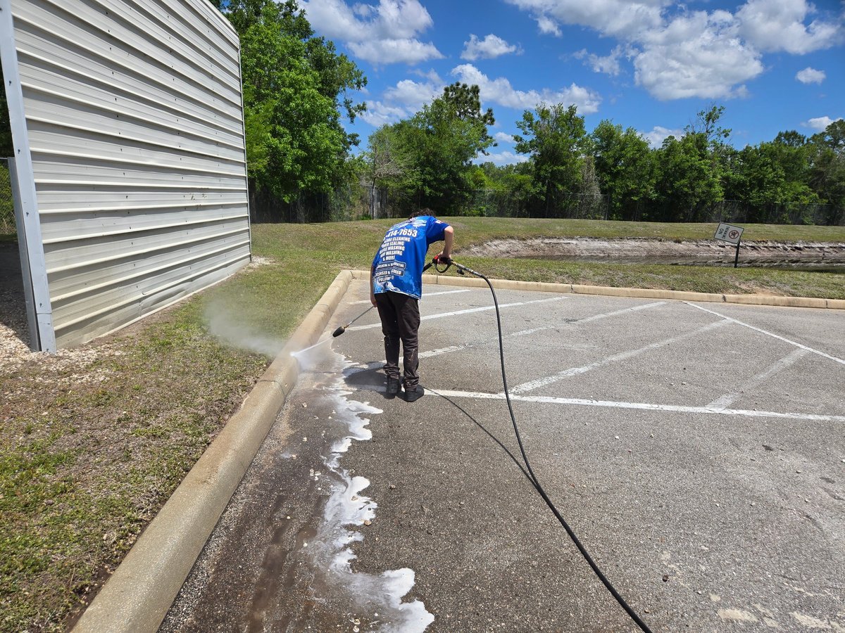 Worker in blue shirt power washing asphalt driveway next to metal building with trees in background
