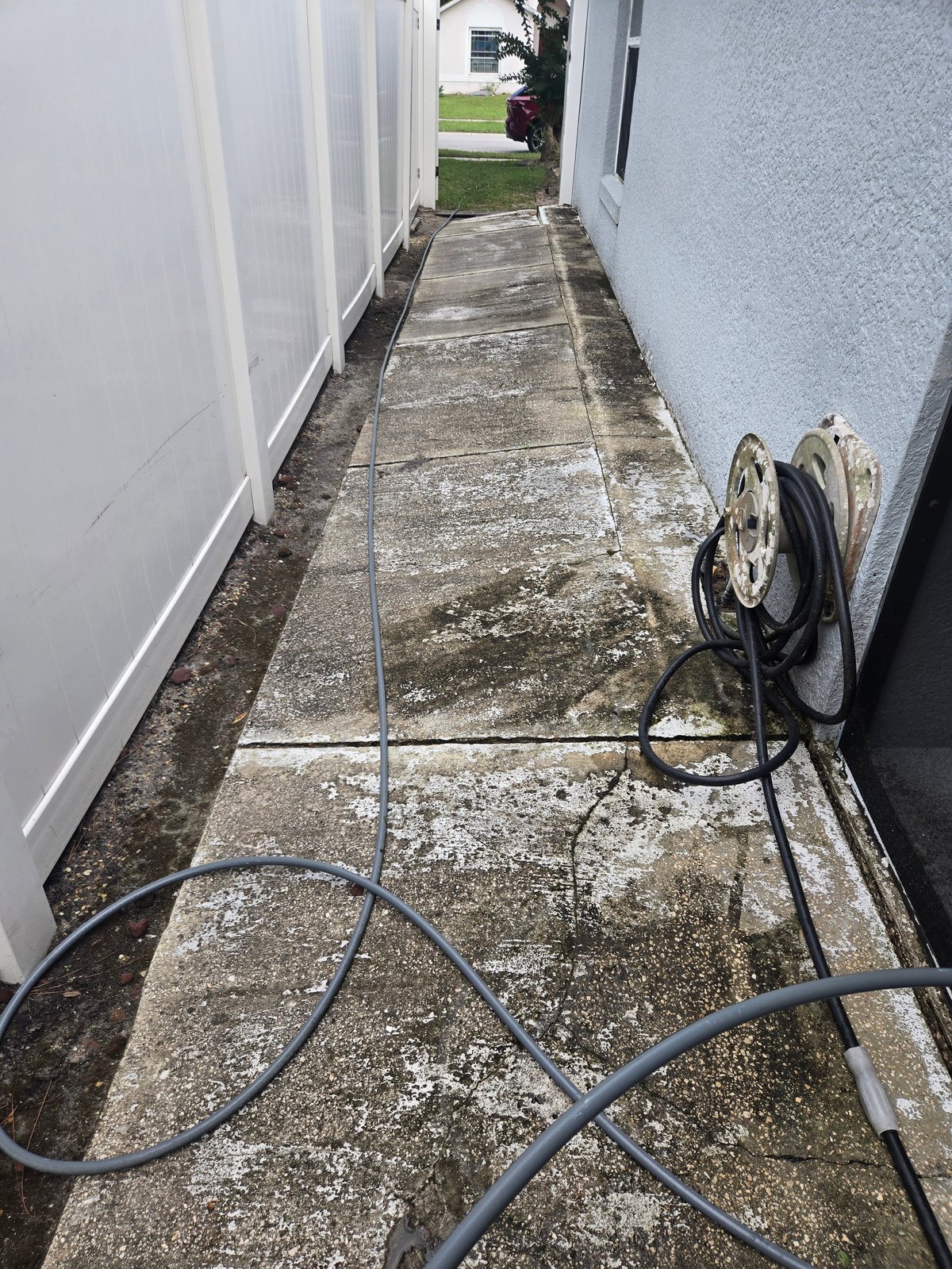 Narrow side yard between two buildings with algae-stained concrete beside a white vinyl fence before cleaning