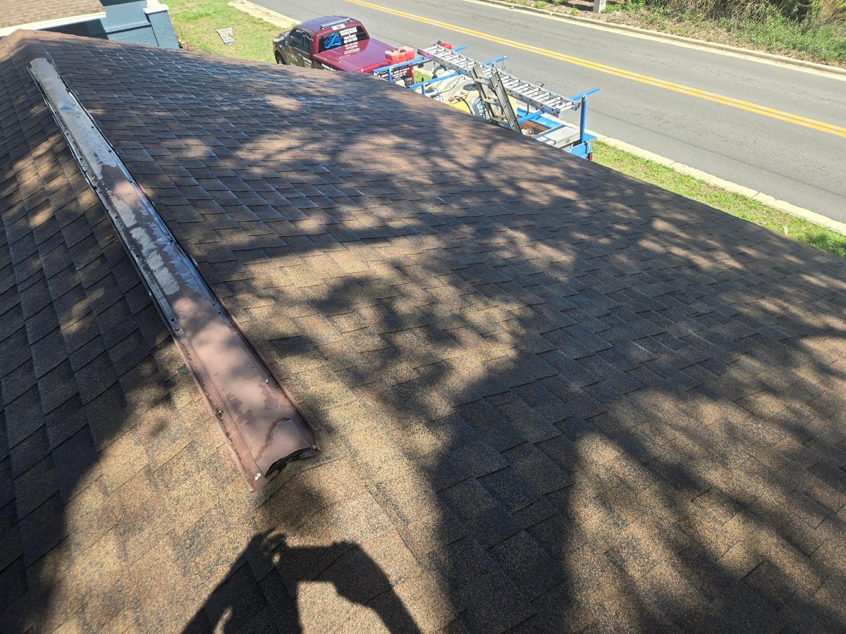 Aerial view of a roof with dark asphalt shingles and tree shadows, with a red truck and utility trailer visible on the road below
