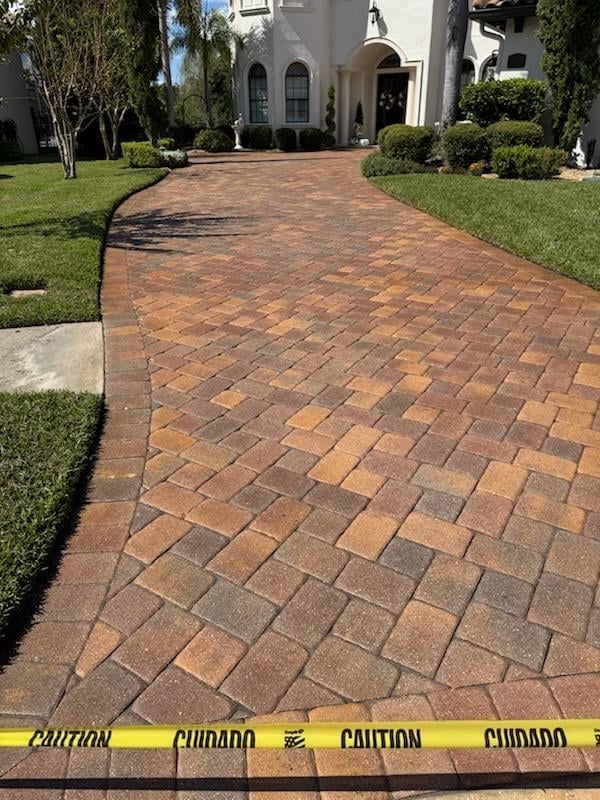 Brick driveway with caution tape leading to white Mediterranean-style home with manicured lawn and landscaping