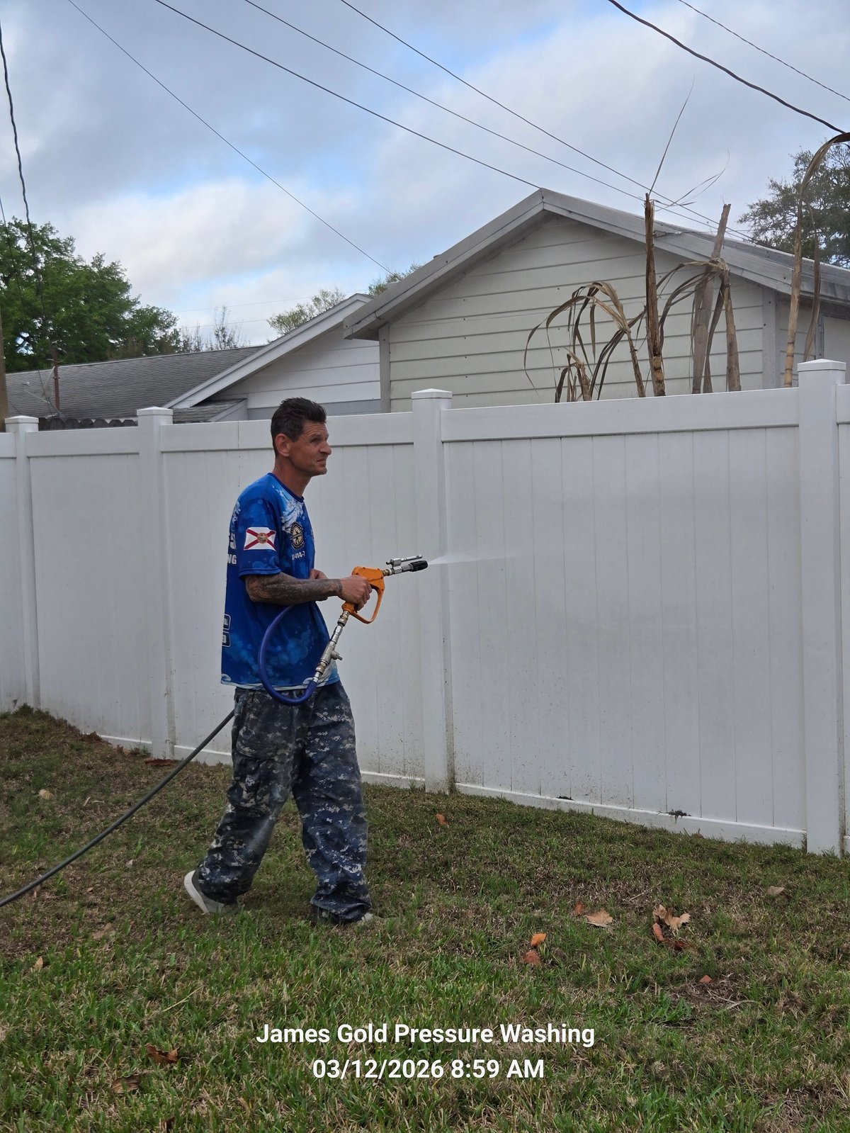 Man in blue shirt and navy pants pressure washing a white vinyl fence in a residential yard