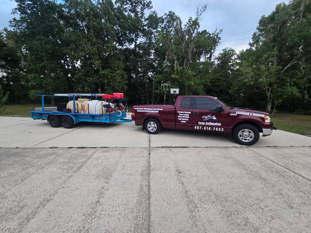 Maroon tow truck parked next to blue flatbed trailer loaded with equipment on a concrete lot with trees in background