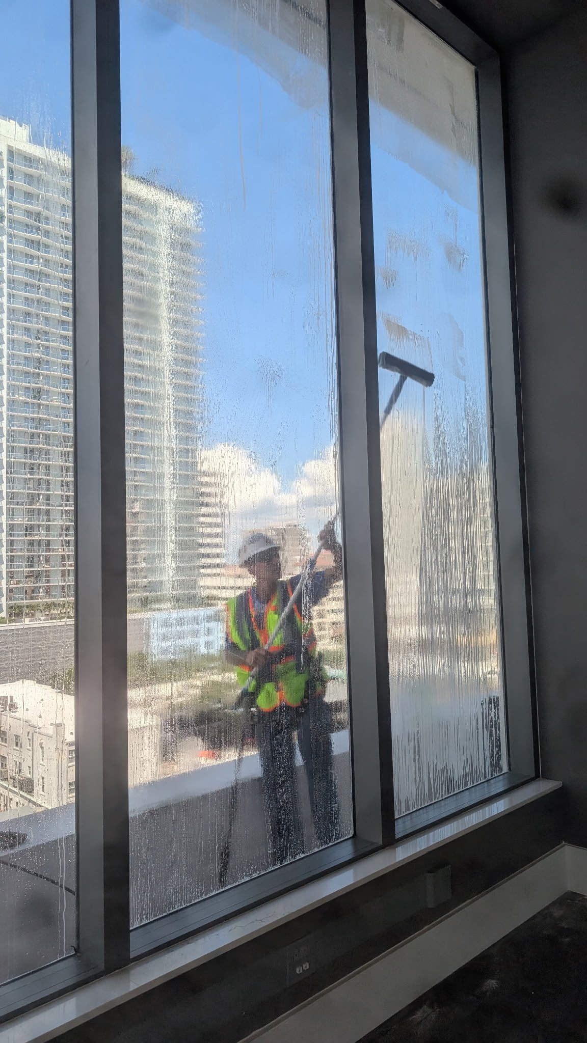 Window cleaner in safety vest cleaning a high-rise building window with squeegee, urban skyline visible in reflection