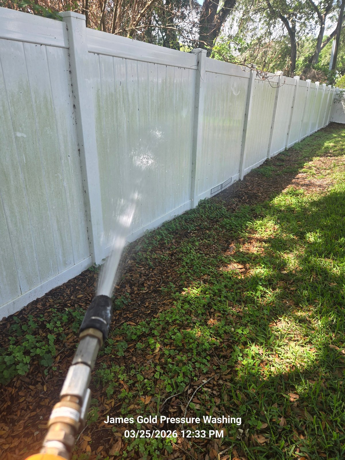White vinyl fence being pressure washed with water hose, showing before and after cleaning with trees in background