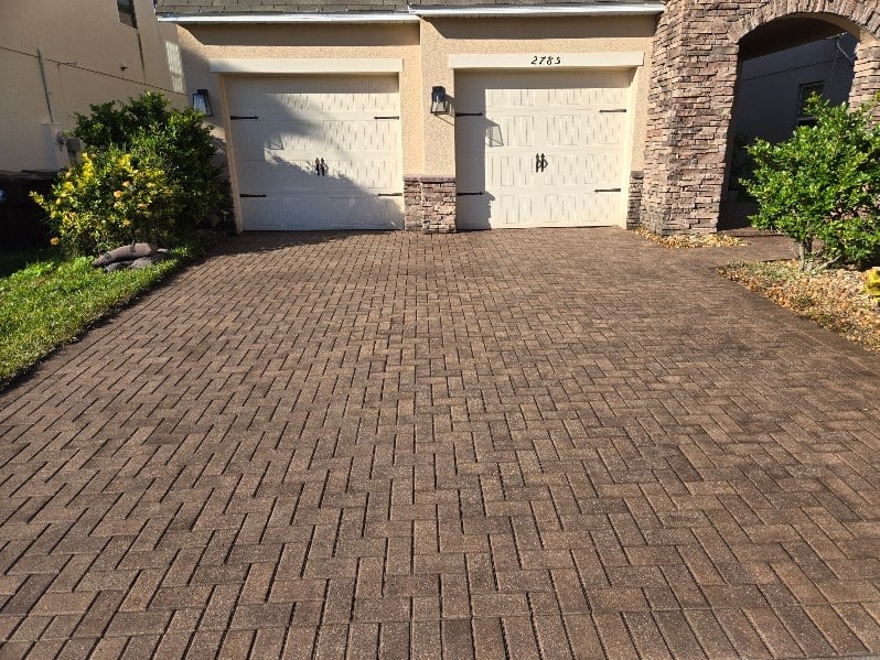 Driveway with two white garage doors, brick paver surface, stone pillars, and landscaping bushes on sides