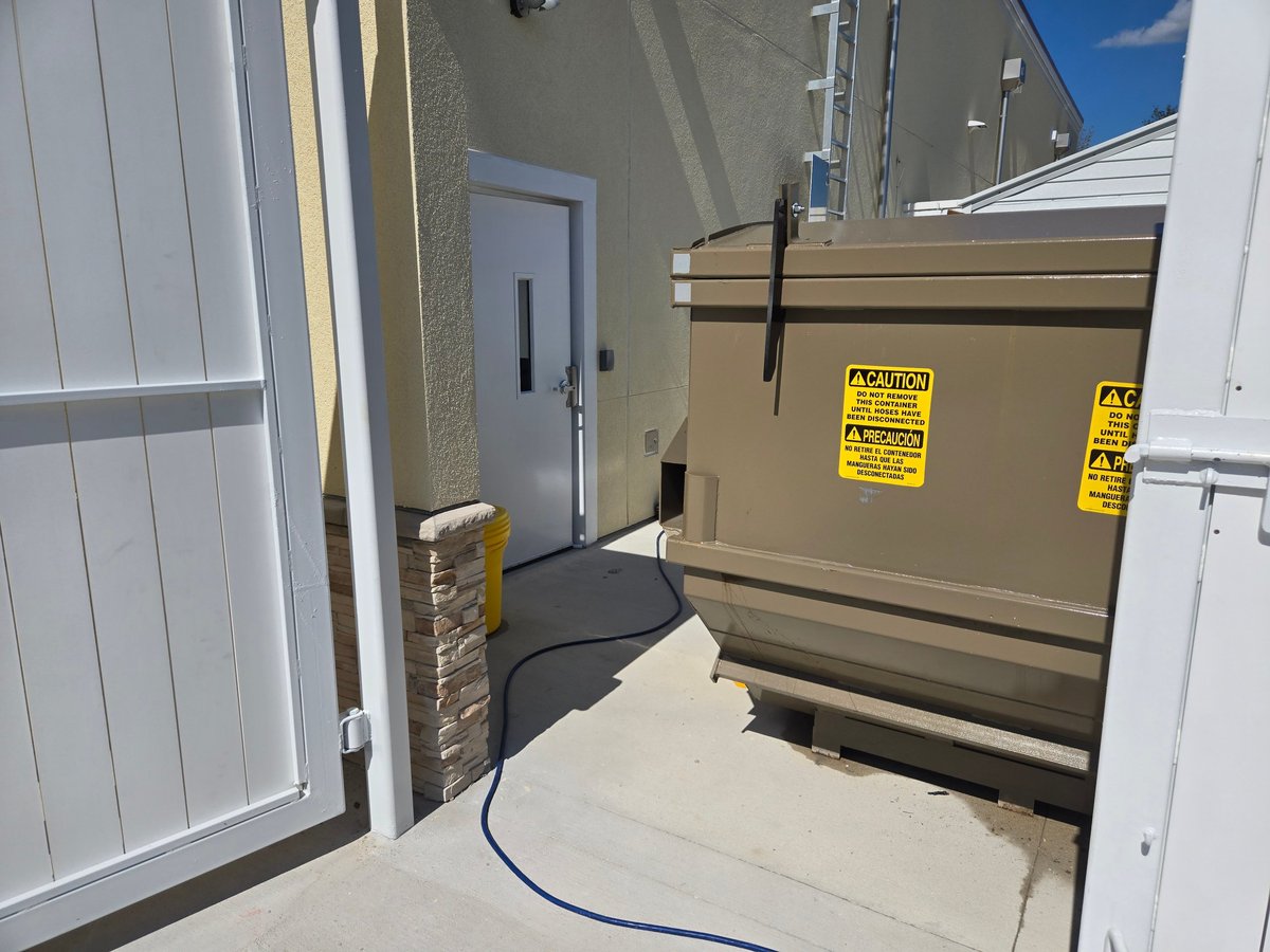 Exterior view of an RV or trailer entrance with white metal doors, a beige storage compartment with yellow warning labels, and a blue hose on concrete ground
