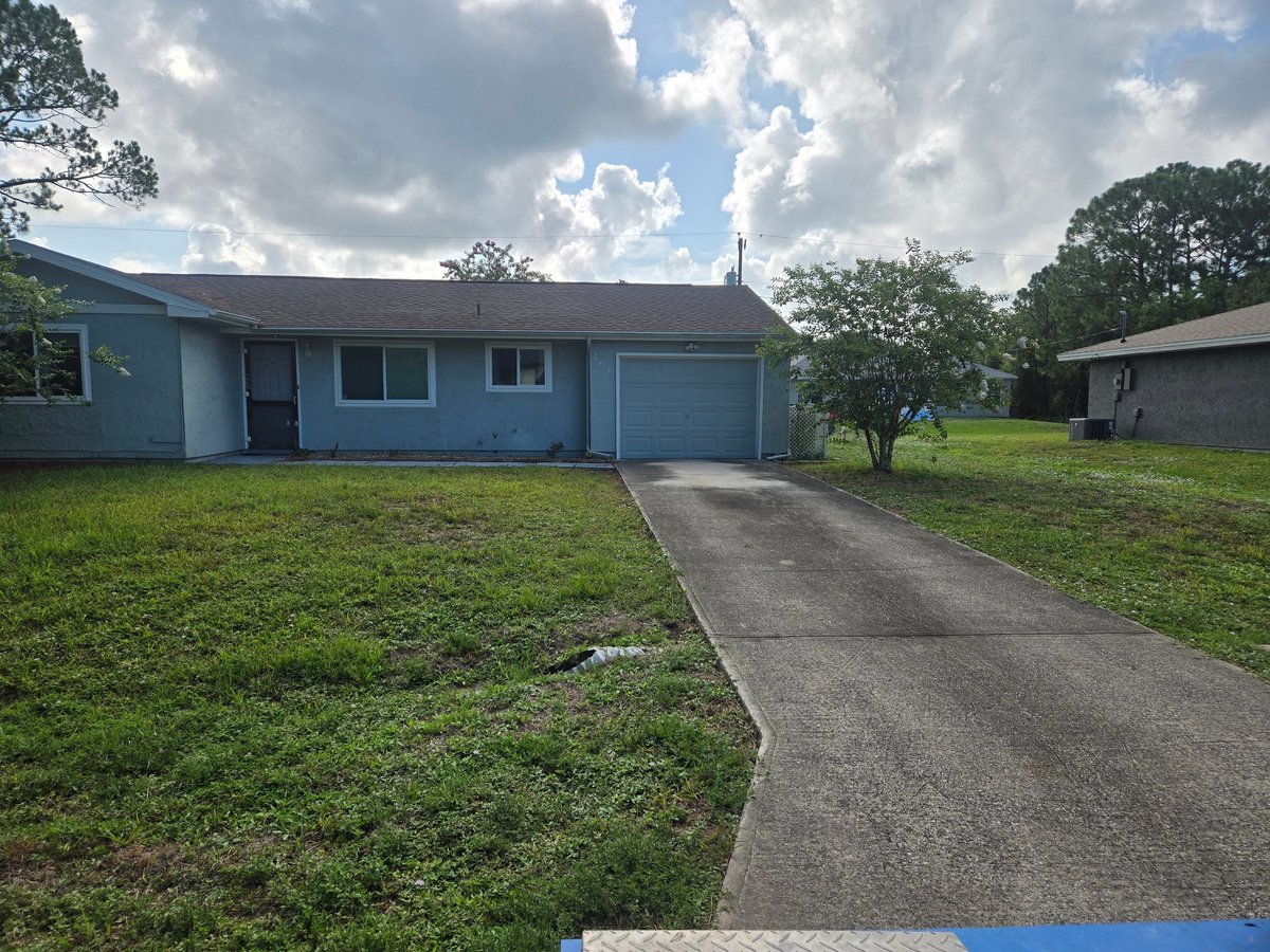 Light blue single-story house with attached garage on a large grassy lot under cloudy skies